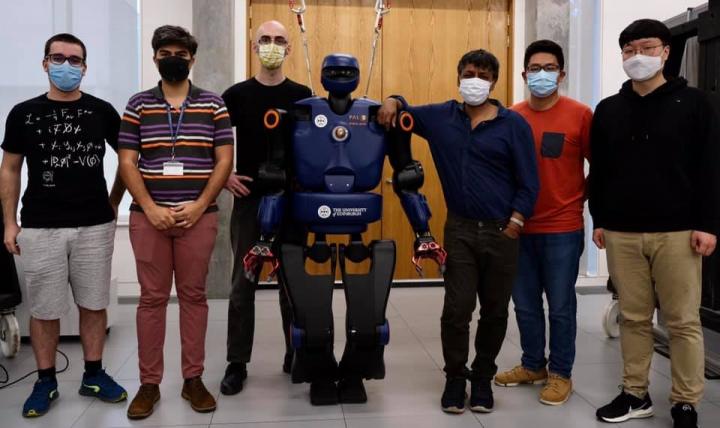 6-foot humaoid robot Talos stands with six members of the robotics research lab, all in masks