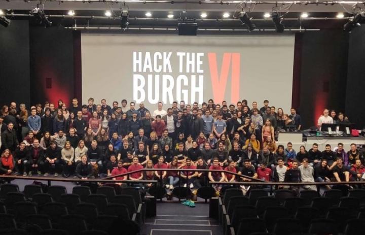 Photo of all the participants of the Hack the Burgh VI, stood on a stage in front of the event logo.